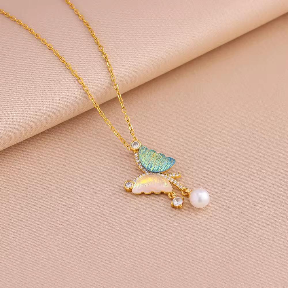 Gold chain necklace with butterfly pendant, blue and pink wings, pearl and crystals, on beige fabric.